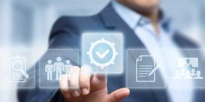 A person in a business suit touches a transparent digital interface showing icons for search, teamwork, settings with a checkmark, and document approval, representing workflow or quality assurance processes, with soft blue lighting and blurred background. Credit: Shutterstock